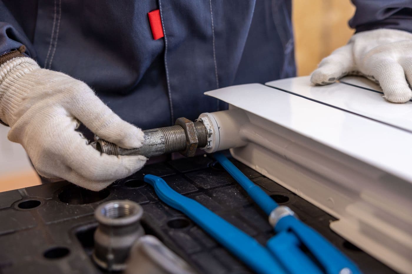 Gloved hands fitting a threaded steel connector to a white radiator, with blue-handled pliers on the workbench.